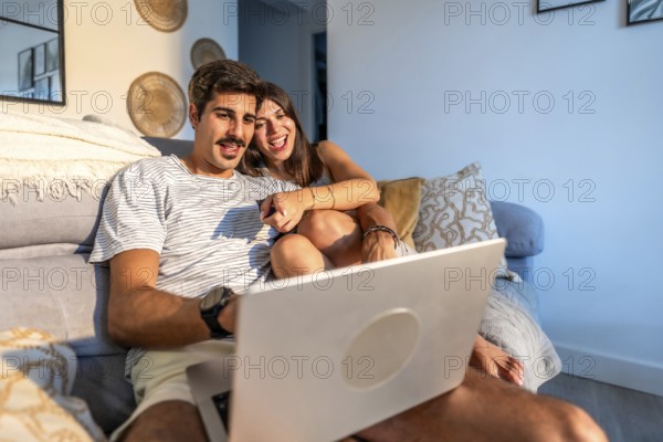 Young couple enjoying a movie on their laptop, laughing and having a good time together on the sofa in their living room