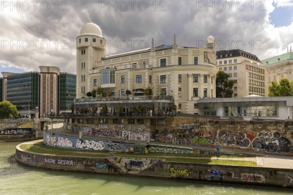A historic building on the riverbank with graffiti on the wall and a cloudy sky Urania Ringstraße Vienna Austria