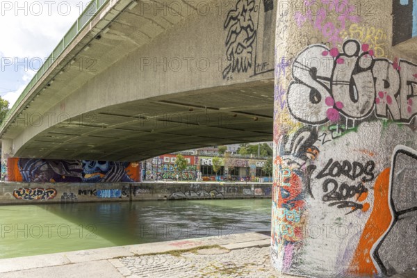 Under a bridge on the river with graffiti-covered columns and riverbank surroundings, Am Donaukanal Innere Stadt Vienna Austria