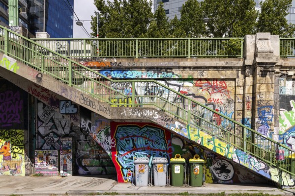 Various graffiti artworks on a concrete wall next to stairs and rubbish bins, Am Donaukanal Innere Stadt Vienna Austria