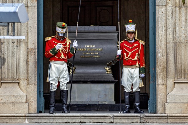 Two uniformed soldiers in red jackets stand guard in front of a marble building, Two soldiers guard a grave in La Paz Bolivia