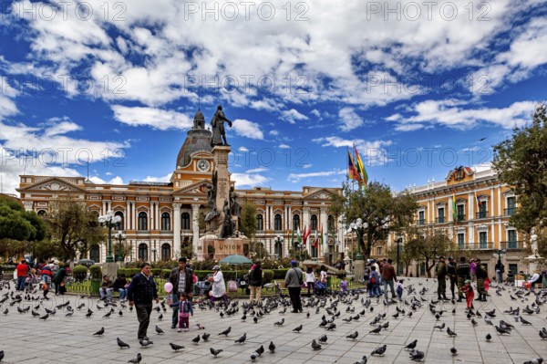 Main square full of people and pigeons in front of a large building with a statue under a blue sky, The presidential palace of La Paz in Bolivia