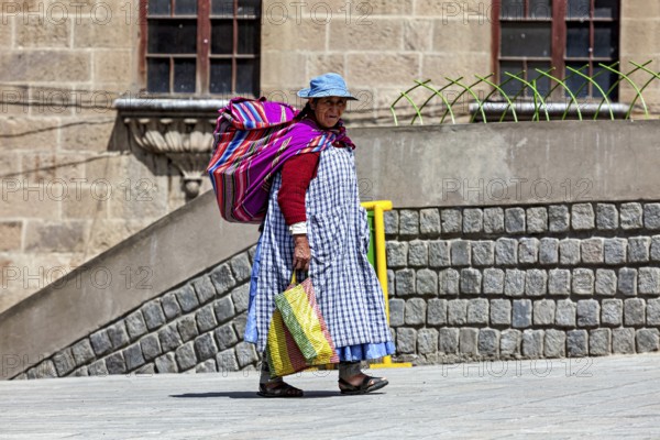 A woman with a colourful bag and traditional clothes walks along a stone wall, Traditionally dressed woman in the streets of La Paz Bolivia