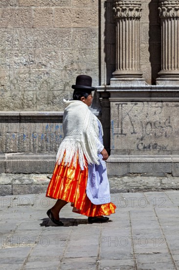 Woman in traditional dress with orange skirt walking along a historical wall, Traditionally dressed woman in the streets of La Paz Bolivia