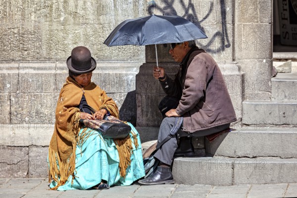 An elderly woman in traditional dress sitting and chatting with a man under an sunshade, A woman and a man with an sunshade sit in La Paz, Bolivia