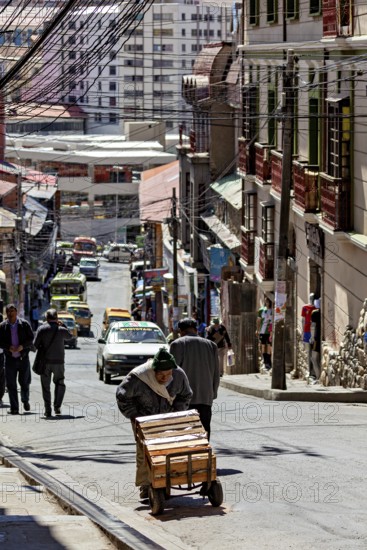 A man pushes a cart on a busy city street. Buildings and cars can be seen in the background, A man pushes a handcart through the streets of La Paz in Bolivia