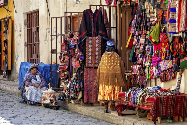 A woman sits in front of a market stall selling a variety of colourful textiles and traditional clothing in a busy street, souvenir shops in La Paz Bolivia