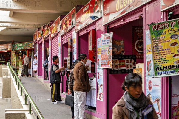 Row of pink market stalls with colourful signs, people ordering food, small shops and restaurants in the market halls of La Paz in Bolivia