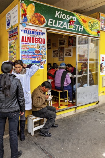 Small food stall with yellow walls, people sitting and eating, menu posters on the wall, Small shops and restaurants in the market halls of La Paz in Bolivia