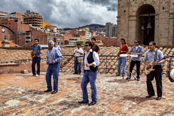 Music group on a city tiled roof in front of an architectural backdrop with a cloudy sky, A music group above the rooftops of La Paz in Bolivia