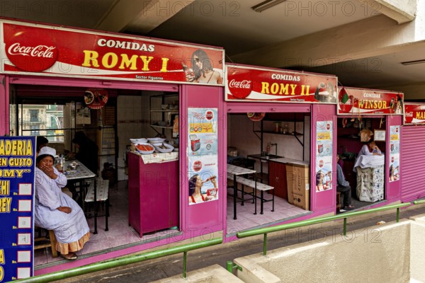 Food stalls with pink awnings and colourful signs, people waiting in a market hall, Small shops and restaurants in the market halls of La Paz in Bolivia