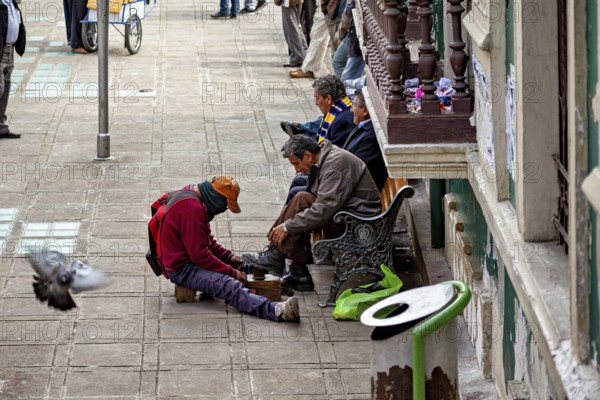 A shoeshine boy shines shoes on a street while passers-by and a pigeon are visible, shoeshine boy in the streets of La Paz in Bolivia