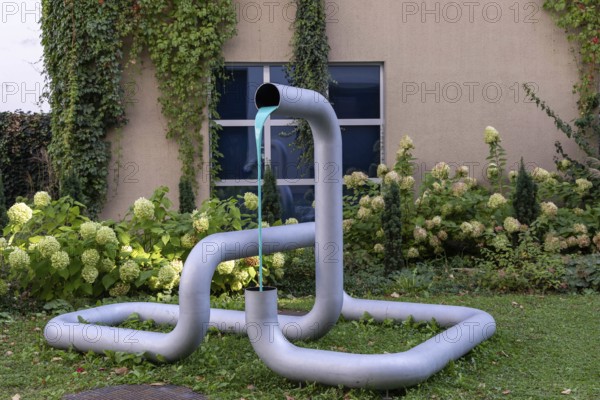 Metal sculpture in the garden with flowing coloured water and plants in the background, city centre Linz Upper Austria Austria