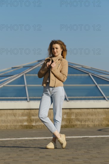 A young woman stands outdoors, wearing a light jacket and jeans. She holds her jacket gently while posing against a backdrop of a sleek glass structure under a clear blue sky