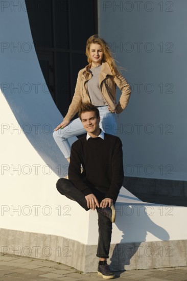 A young man sits on a low wall with his leg bent while a woman stands above him, both smiling. The scene captures a moment of joy in a bright location showcasing modern architectural design