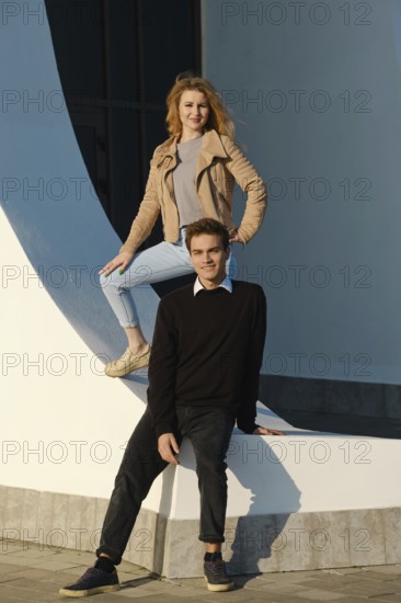 A young couple enjoys a sunny day in an urban area, posing creatively at a striking architectural structure. The female stands on the edge while the male sits below, showcasing their playful spirit