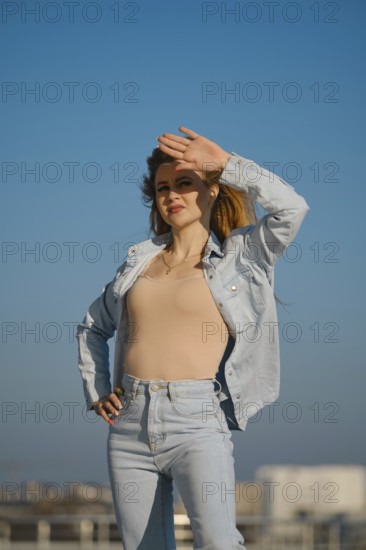 A woman stands outdoors on a sunny day, shielding her eyes from the sunlight. She wears a light denim jacket and jeans, with a confident expression, creating a relaxed atmosphere