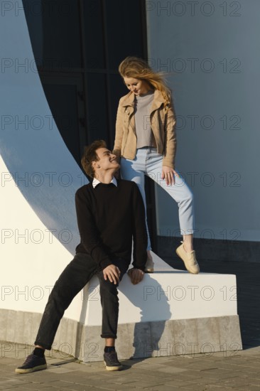 Two young adults share a light-hearted moment by a large curved structure in an urban setting. The sun shines brightly as they engage in playful interaction, creating a joyful atmosphere