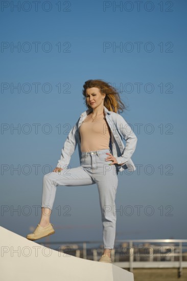 A young woman stands on a ledge outdoors, wearing light denim and looking directly at the camera. Her hair flows in the breeze, highlighting a sunny day with a bright blue sky