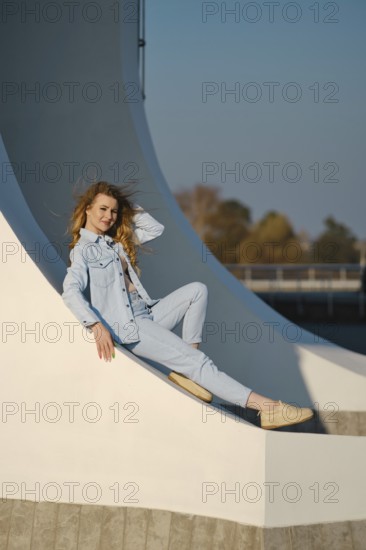 A woman with long hair is sitting comfortably on a curved architectural element. She wears a light denim outfit and smiles as sunlight highlights her pose