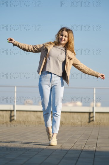 A young woman stands on one foot with arms outstretched, smiling under a clear blue sky. She wears a light fleece jacket and jeans, enjoying a sunny day at a city location with a railing in view