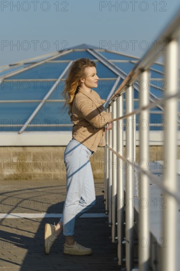 A young woman stands by the observation deck, leaning on a white railing. She gazes out at the clear blue sky, enjoying the warm sunlight during a calm day