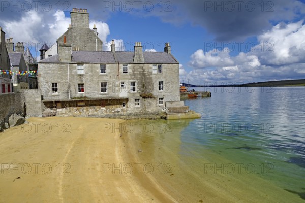 Old stone cottage on a quiet beach with clear water and partly cloudy skies, Lerwick, Shetland Islands, Scotland, United Kingdom