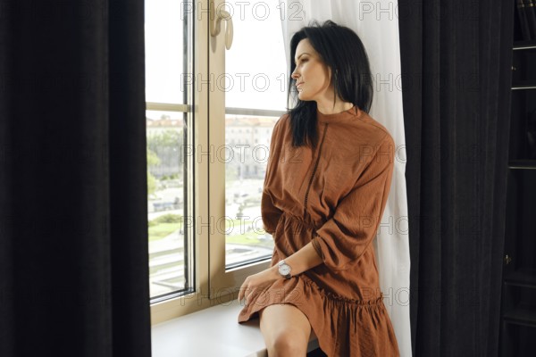 A woman in a brown dress relaxes by a large window, gazing thoughtfully outside. Sunlight filters through the curtains, illuminating her peaceful expression and the vibrant cityscape beyond