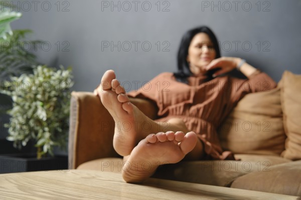 A barefoot woman sits comfortably on a cozy couch, her bare feet resting on a wooden table. The modern living room features neutral tones and indoor plants, providing a relaxed atmosphere