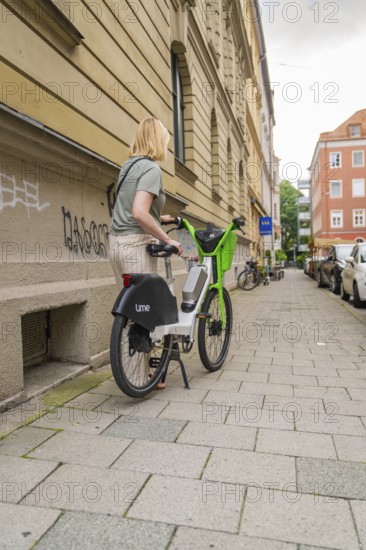 Woman pushing a green e-bike along a city street, graffiti on the wall, Munich, Germany
