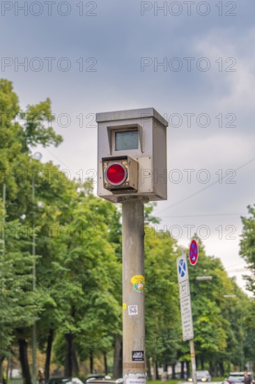Traffic surveillance camera on a pole, surrounded by trees and road signs under a cloudy sky, Munich, Germany