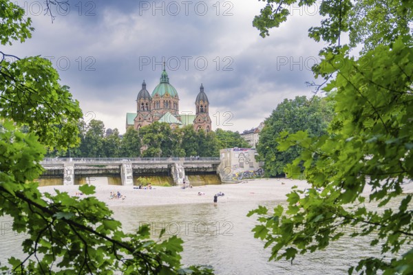 Imposing church behind a river, lined with trees and sandbanks under a cloudy sky, Munich, Germany