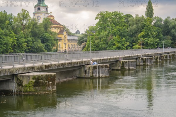 View of a bridge over a quiet river, surrounded by green trees and an impressive building in the background, Munich, Germany
