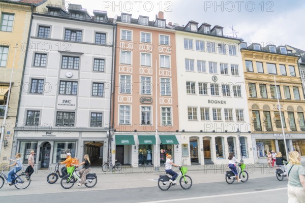 Passers-by ride along on bicycles in front of colourful building facades in Munich, Munich, Germany