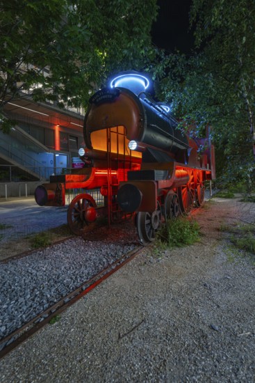 Luminous steam engine as an art installation surrounded by trees at night, Munich, Germany