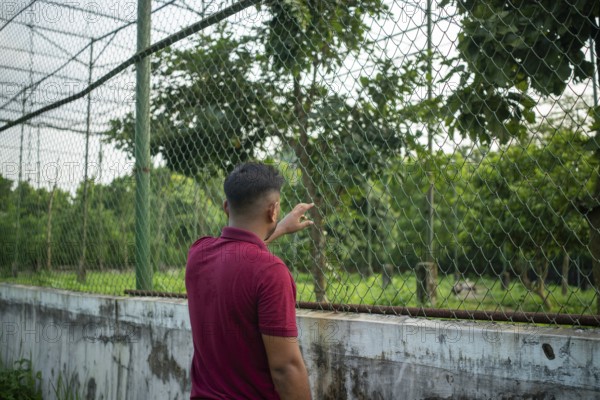 A man stands by a chain-link fence surrounded by lush greenery, appearing contemplative-Dhaka, Bangladesh