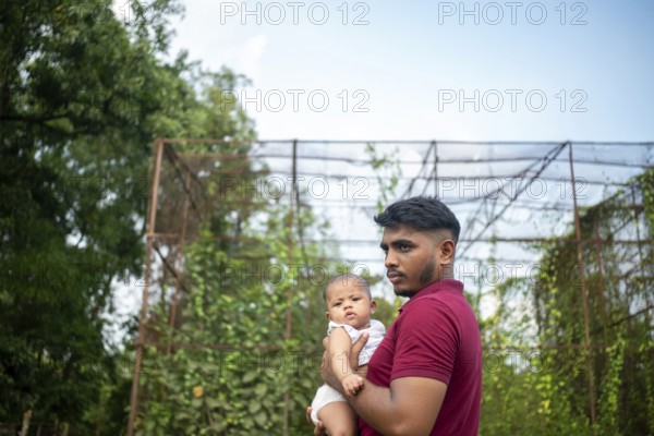 A man holds a baby outdoors by a metal structure with greenery in a relaxed setting-Dhaka, Bangladesh