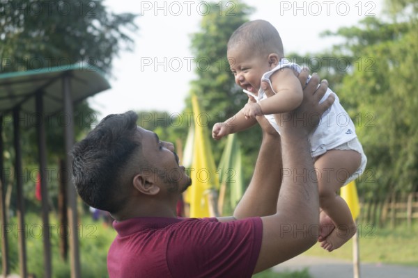 A man joyfully lifts a smiling baby in an outdoor setting-Dhaka, Bangladesh