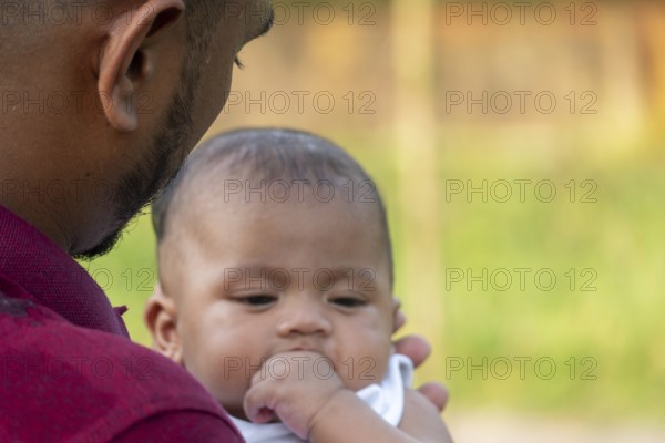Baby calmly looks down, held by a gentle man outdoors-Dhaka, Bangladesh