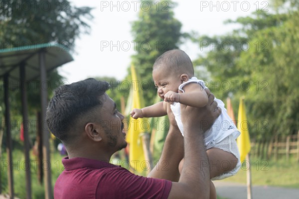 A parent is lifting a baby, creating a joyful moment in a green outdoor setting-Dhaka, Bangladesh