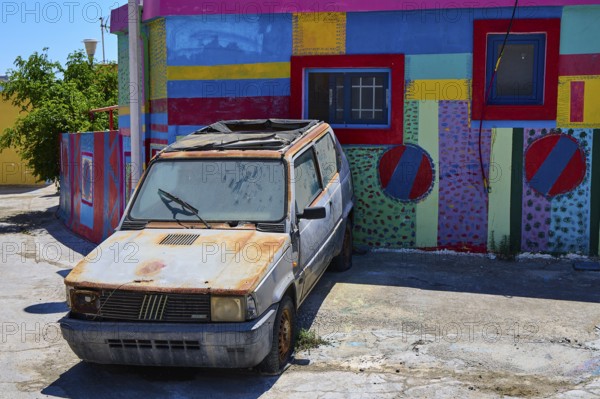 Neglected old car in front of a colourfully painted urban building, Kattavia village, southern tip of Rhodes, Rhodes, Dodecanese, Greek Islands, Greece