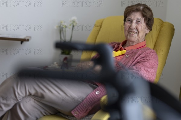 90+ year old senior citizen sitting laughing in her room in a retirement home behind a walker in a yellow armchair, Cologne, North Rhine-Westphalia, Germany