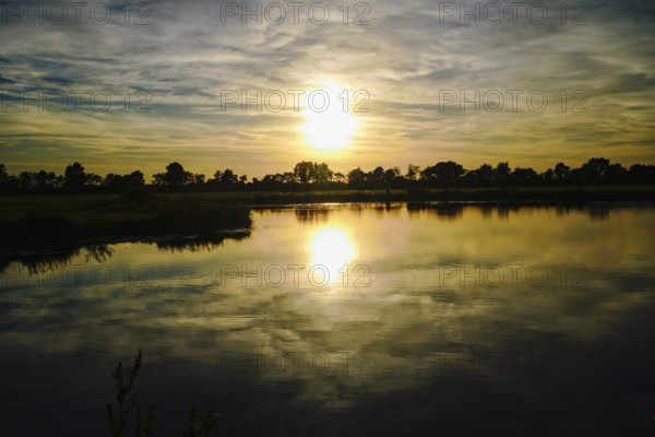 The setting sun paints the sky in warm colours that are reflected in the lake, natural landscape in Wüsting, 27798 Hude, Lower Saxony, Germany