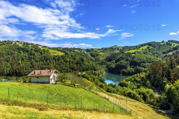 Surrounded by green rolling hills and lush forests, the tranquil ibiur reservoir features a charming house nestled on a hillside under a vibrant blue sky with fluffy clouds