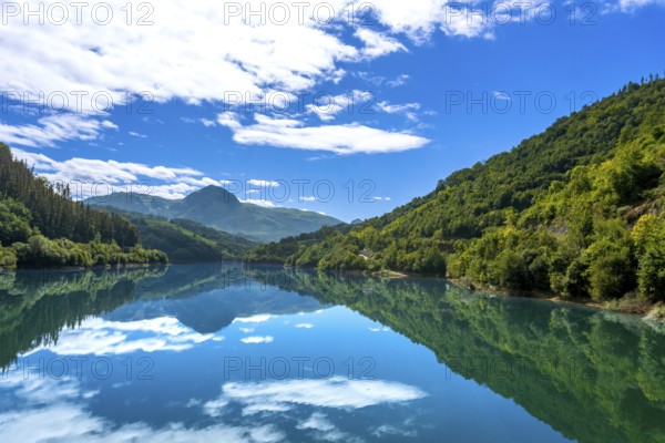 The tranquil waters of ibiur reservoir mirror the vibrant blue sky and fluffy white clouds, creating a picturesque scene surrounded by the verdant slopes of the basque mountains in tolosa, gipuzkoa