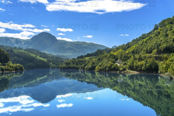 The serene ibiur reservoir in tolosa, gipuzkoa, reflects the surrounding mountains and blue sky with white clouds on a sunny day, creating a tranquil scene in the heart of the basque country