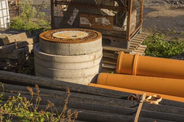 Borken, NRW, Germany, Orange and black pipes, concrete rings, and a rusty manhole cover sit at a construction site, awaiting installation in a sewer system