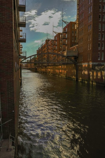 Sunlight is reflected in the water of the canal between tall brick buildings. View from the Wilhelminenbrücke over the Kehrwiederfleet towards the Speicherstadt, 20457 Hamburg, Germany