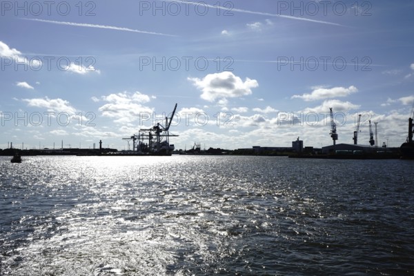 An industrial harbour with cranes and reflecting sunlight on the water under a blue sky. The view towards the Hansa harbour, seen from the Norderelbe in 20457 Hamburg