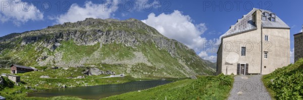 Panoramic photo view to the right left side of Ospizio Hospiz Herberge San Gottardo St. Gotthard built in the 13th century from 1230 with small entrance door main entrance to chapel Cappella di San Gottardo on pass summit of Gotthard Pass, left mountain lake Lago di Monti, Swiss Alps, Airolo, Canton Ticino, Ticino, Switzerland